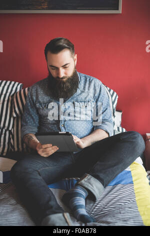 Jeune homme barbu using digital tablet on bed Banque D'Images