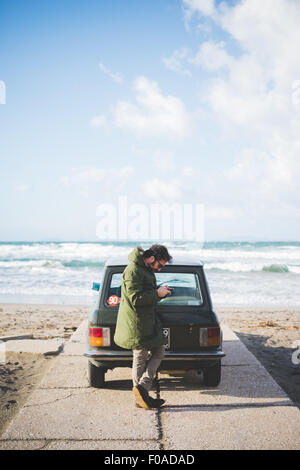 Homme avec vintage voiture garée sur la lecture de textes, plage smartphone Sorso, Sassari, Sardaigne, Italie Banque D'Images