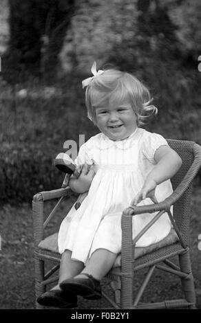 Une petite fille dans une robe assis sur une chaise avec une brosse et souriant à la caméra. Banque D'Images