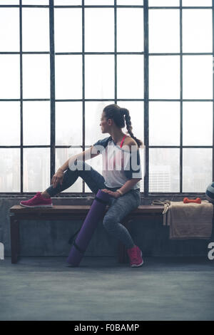 Une femme assise, perdue dans le buzz de la ville ci-dessous, et se détendre après avoir eu un bon entraînement. Une jambe au repos sur le banc, et tenant son tapis de yoga avec son autre main, elle est paisible. Banque D'Images