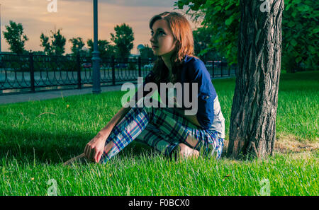 Jeune rousse boho girl sitting on grass rétro-éclairé et à la voiture. Coucher du soleil la lumière colorée. Banque D'Images