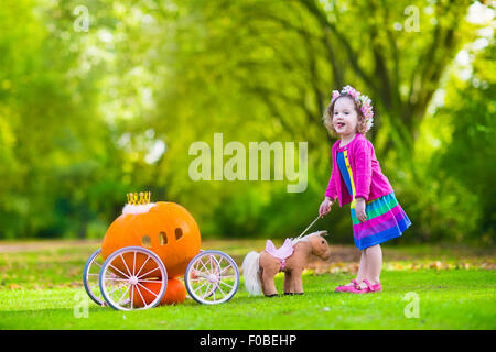 Cute little girl playing bouclés conte Cendrillon holding baguette magique à côté d'un chariot de citrouille en automne park à l'Halloween. Banque D'Images