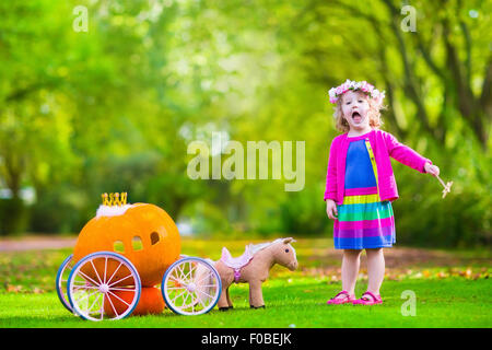 Cute little girl playing bouclés conte Cendrillon holding baguette magique à côté d'un chariot de citrouille en automne park à l'Halloween. Banque D'Images