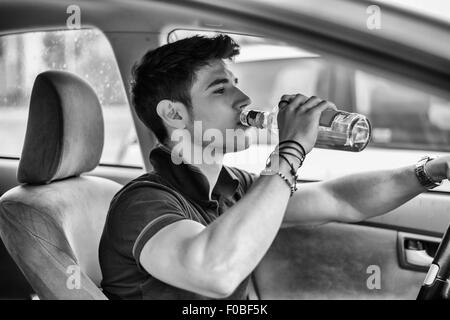 Beau jeune homme conduisant sa voiture tandis que boire de l'alcool dans la circulation. Photo en noir et blanc Banque D'Images