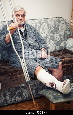 Senior man with broken leg sitting on sofa at home, tenant des béquilles Banque D'Images