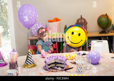 Deux filles assis à table d'anniversaire avec gâteau jouant avec smiley face balloon Banque D'Images