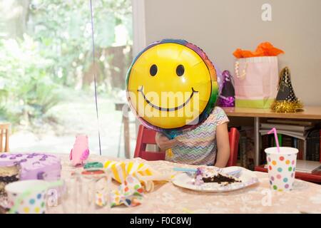 Fille assise à la table d'anniversaire avec gâteau jouant avec smiley face balloon Banque D'Images