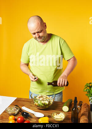 Salade fraîche ingrédients sur la table, homme d'âge moyen verse de l'huile - fond jaune Banque D'Images