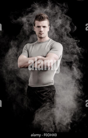 Portrait de beau jeune homme robuste en t-shirt gris sur fond noir, avec les bras croisés sur la poitrine, looking at camera. Fumée secondaire b Banque D'Images