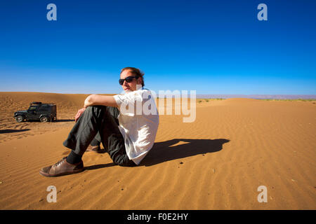 Jeune homme assis sur une dune de sable à Merzouga et regardant la caméra, à l'arrière-plan, il y a un 4x4 véhicule stationné. Banque D'Images