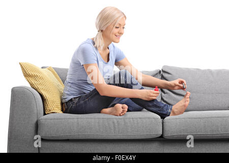 Jeune femme blonde avec un polissage ongles son vernis à ongles rouge assis sur un canapé gris isolé sur fond blanc Banque D'Images