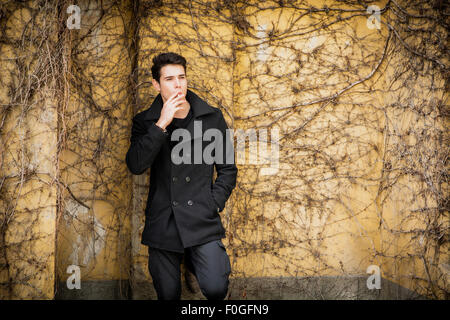 Handsome young man standing contre un mur couvert de plantes, porter des vêtements d'hiver, fumer cigarette Banque D'Images