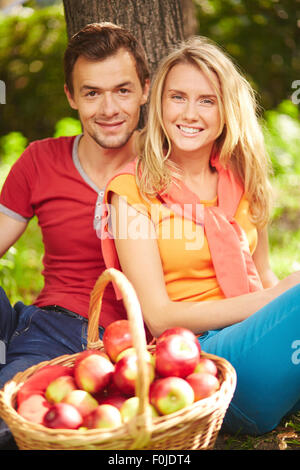 Beau couple avec panier rempli de pommes rouges assis par tree Banque D'Images