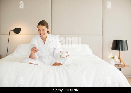 Smiling woman in bathrobe drinking coffee and texting on cell phone on bed Banque D'Images