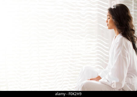 Serene woman in bathrobe méditant à fenêtre ensoleillée Banque D'Images