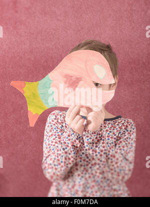 Portrait de petite fille en tenue d'organiser une coupe dessin d'un poisson en face de son visage. Elle est timide et se cacher derrière elle. Banque D'Images