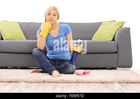 Jeune femme avec des gants de nettoyage tenant une bouteille de jet et assis sur le sol en face d'un canapé gris Banque D'Images