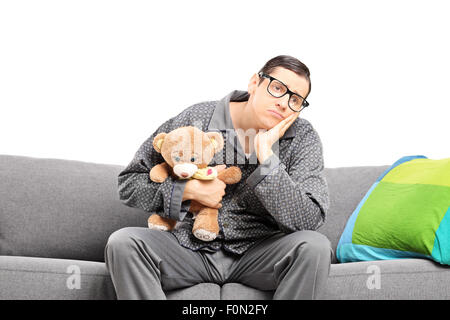 Homme triste dans pajamas holding a teddy bear assis sur un canapé isolated on white Banque D'Images