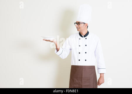 Portrait of handsome male chef indien en uniforme présentant un plat vide et souriant, debout sur fond uni avec l'ombre, Banque D'Images