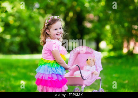 Petite fille poussant poussette jouet avec l'ours. Tout-petit enfant en robe rose jouant à la poupée buggy. Kids Birthday party. Banque D'Images