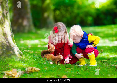 Kids feeding écureuil dans le parc de l'automne. Petit garçon et fille en robe rouge et des bottes de pluie regarder les animaux sauvages dans la forêt d'automne Banque D'Images