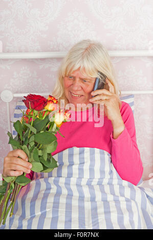 Femme heureuse en pyjama rose assis au lit avec un bouquet de roses et à parler au téléphone Banque D'Images
