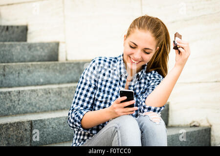 Adolescent - jeune femme de manger du chocolat en rue et à la recherche dans le téléphone Banque D'Images