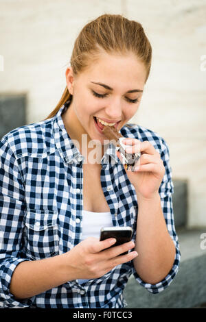 Adolescent - jeune femme de manger du chocolat en rue et à la recherche dans le téléphone Banque D'Images