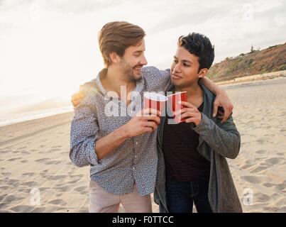 Deux amis de sexe masculin ayant un verre ensemble à la plage, les bras autour de l'autre Banque D'Images