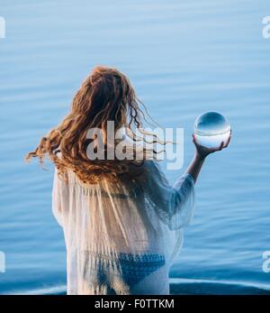 Vue arrière du jeune femme aux longs cheveux rouges dans le lac permanent holding crystal ball Banque D'Images