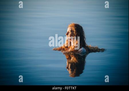 Tête et épaules de jeune femme aux longs cheveux rouges dans le lac avec les yeux fermé Banque D'Images