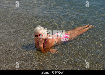 Portrait de senior lady c'est couché dans l'eau peu profonde de la mer de galets de plage. Banque D'Images