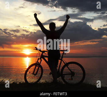 Silhouette d'un homme avec un vélo de montagne sur la rive du fleuve au coucher du soleil Banque D'Images