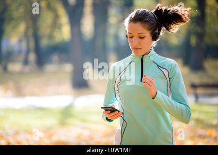 Jeune femme avec un casque jogging en automne nature et à la recherche de téléphone mobile Banque D'Images