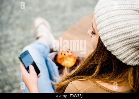 Ado - young woman eating muffin dans la rue et à la recherche dans le téléphone Banque D'Images