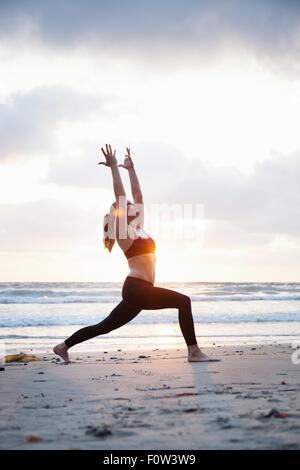 Mid adult woman practicing warrior yoga pose sur la plage au coucher du soleil Banque D'Images