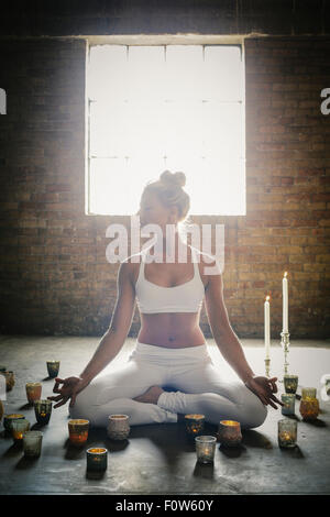 Une femme blonde dans un blanc petit top et leggings, assise sur le sol entourée de bougies, faisant du yoga. Banque D'Images