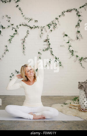 A smiling woman sur un tapis blanc dans une chambre, s'étirant les bras. Une plante rampante sur le mur derrière elle. Banque D'Images