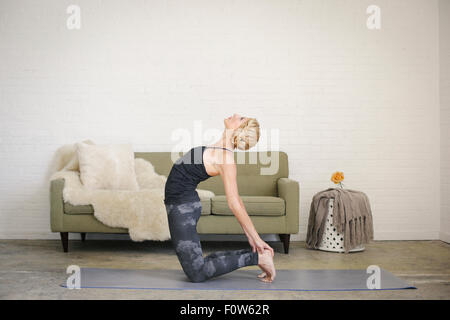 Une femme blonde à genoux sur un tapis de yoga dans une salle, faire du yoga, se pencher en arrière avec ses mains toucher ses talons. Banque D'Images