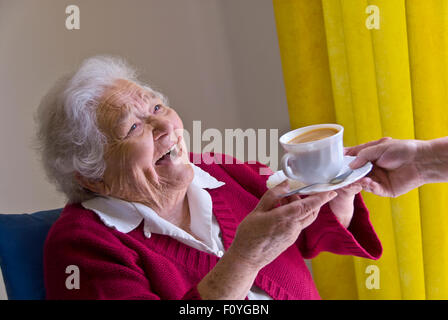 Personnes âgées 90 HEUREUX SOINS THÉ ACCUEIL SOIGNANT VISITEZ heureux rire dame âgée recevant une tasse de thé d'infirmière soignant compagnon dans le salon spacieux lumineux Banque D'Images