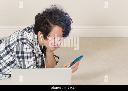 Smiling teen girl looking at cell phone avec ordinateur en avant-garde tout en étant allongé à écouter de la musique à la maison. Banque D'Images