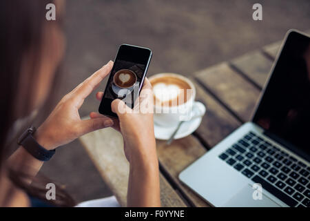 Femme de prendre une photo d'une tasse de café avec son téléphone intelligent tout en restant assis dans un café. Banque D'Images