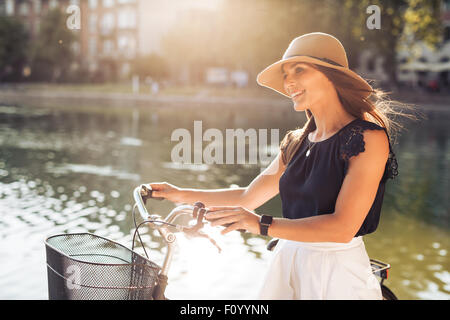 Image de belle femme avec un vélo à l'heure d'été près de l'étang au parc. Modèle féminin wearing hat à la voiture en souriant. Banque D'Images