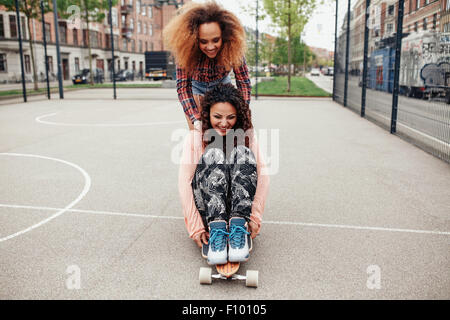 Femme poussant son ami sur longboard. Les filles en patinage de Hipster un terrain de basket dans la ville. Banque D'Images