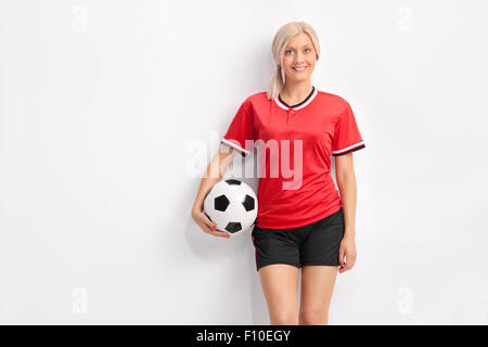 Jeune joueur de soccer féminin dans un maillot rouge tenant une balle et appuyé contre un mur Banque D'Images