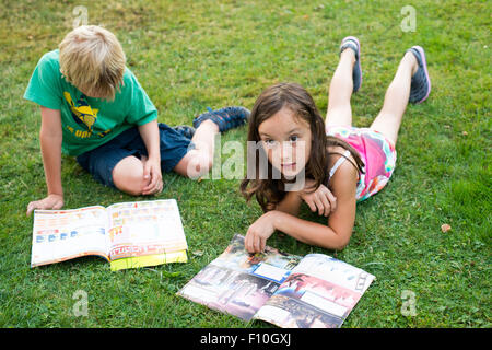 Lecture enfants jouissant ensemble et la lecture d'un magazine portant sur l'herbe en plein air Banque D'Images