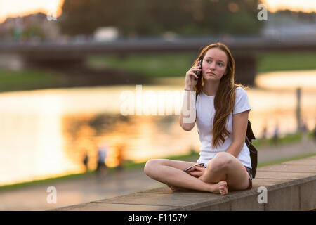 Jeune fille assise sur le bord de l'eau pendant le coucher du soleil et parle sur un téléphone mobile. Banque D'Images