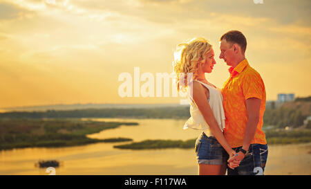 Couple d'amoureux se tenant la main et se regarder dans les yeux sur la rive. Main dans la main. Le coucher du soleil. Journée d'été. Banque D'Images