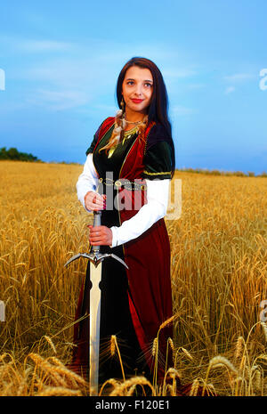 Souriante Jeune femme avec robe d'ornement et de l'épée à la main debout sur un champ de blé avec le coucher du soleil. Fond naturel.. Banque D'Images