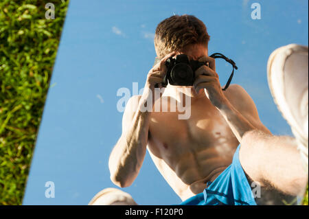Mais pauvre homme musclé avec son top off prendre des photos de lui-même dans un miroir. Résumé photos. Le miroir est allongé sur l'herbe. Banque D'Images